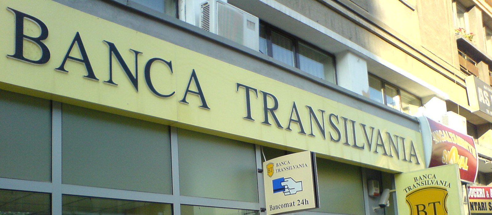 Firma luminoasă Banca Transilvania pe fațada unei sucursale, cu panoul pentru bancomat non-stop dedesubt